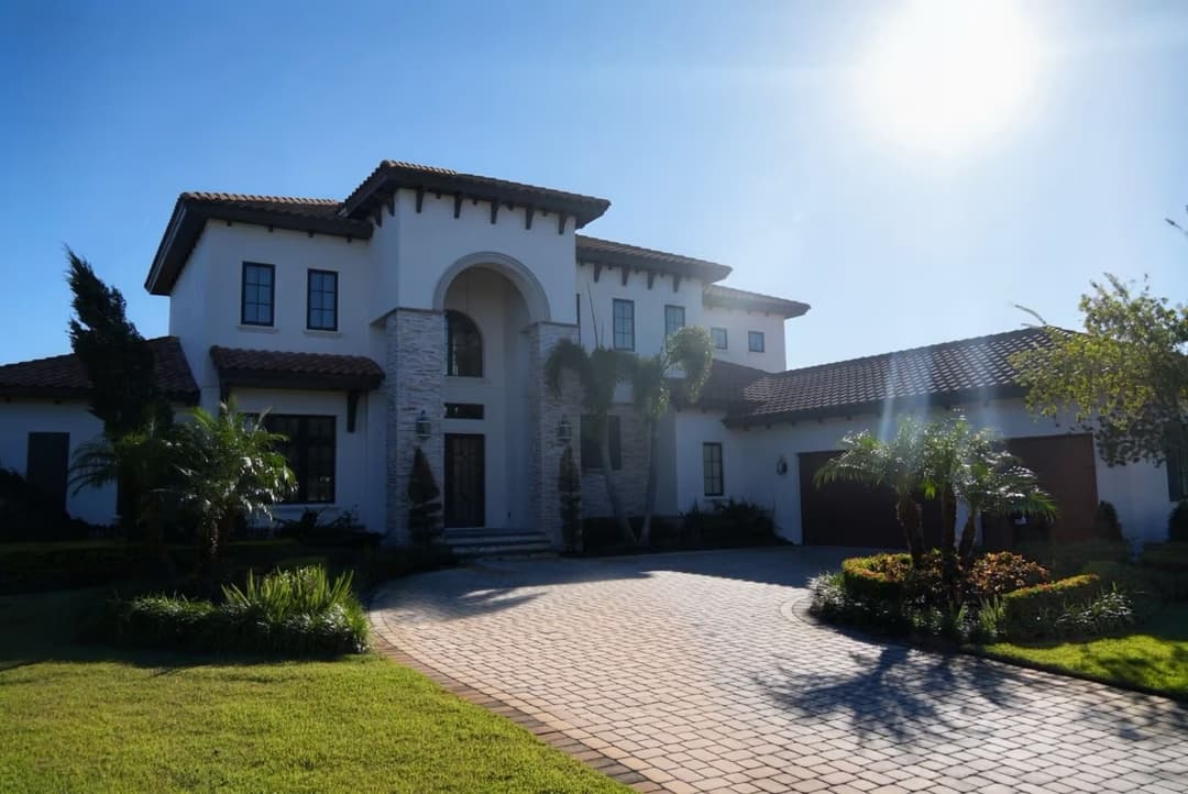 Modern luxury home with palm trees and brick driveway under bright sunlight.