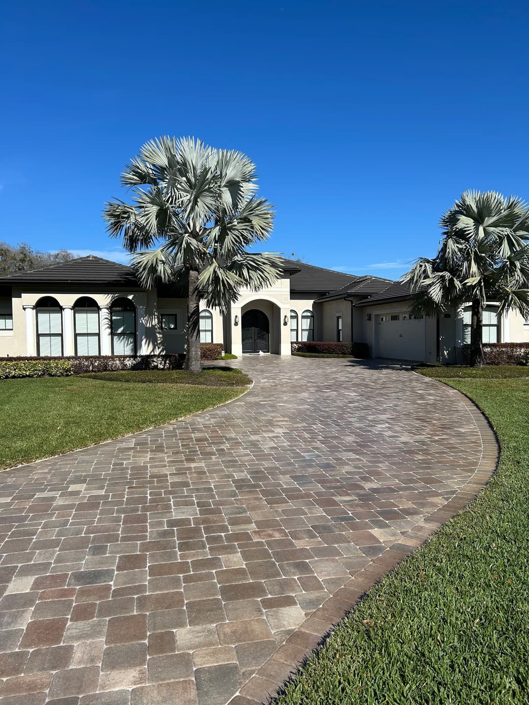 Modern home with palm trees and paver driveway under a clear blue sky.