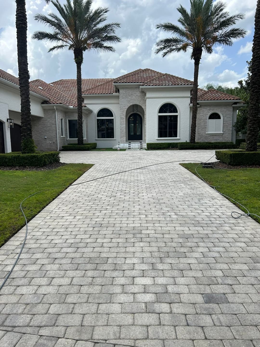 Luxury home entrance with paver driveway and palm trees under a cloudy sky.