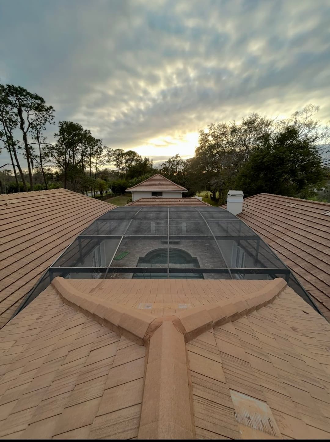 Aerial view of a home's roof with a pool enclosure at sunset surrounded by trees.