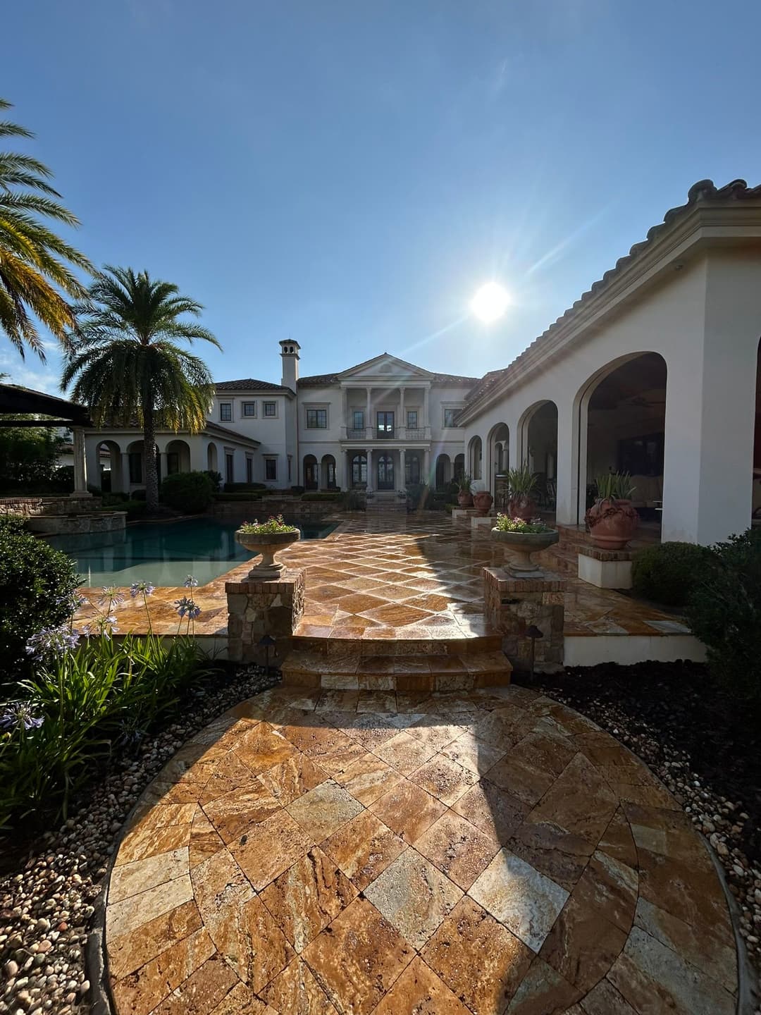 Luxury mansion with palm trees and pool, featuring a stone pathway and sunny sky.