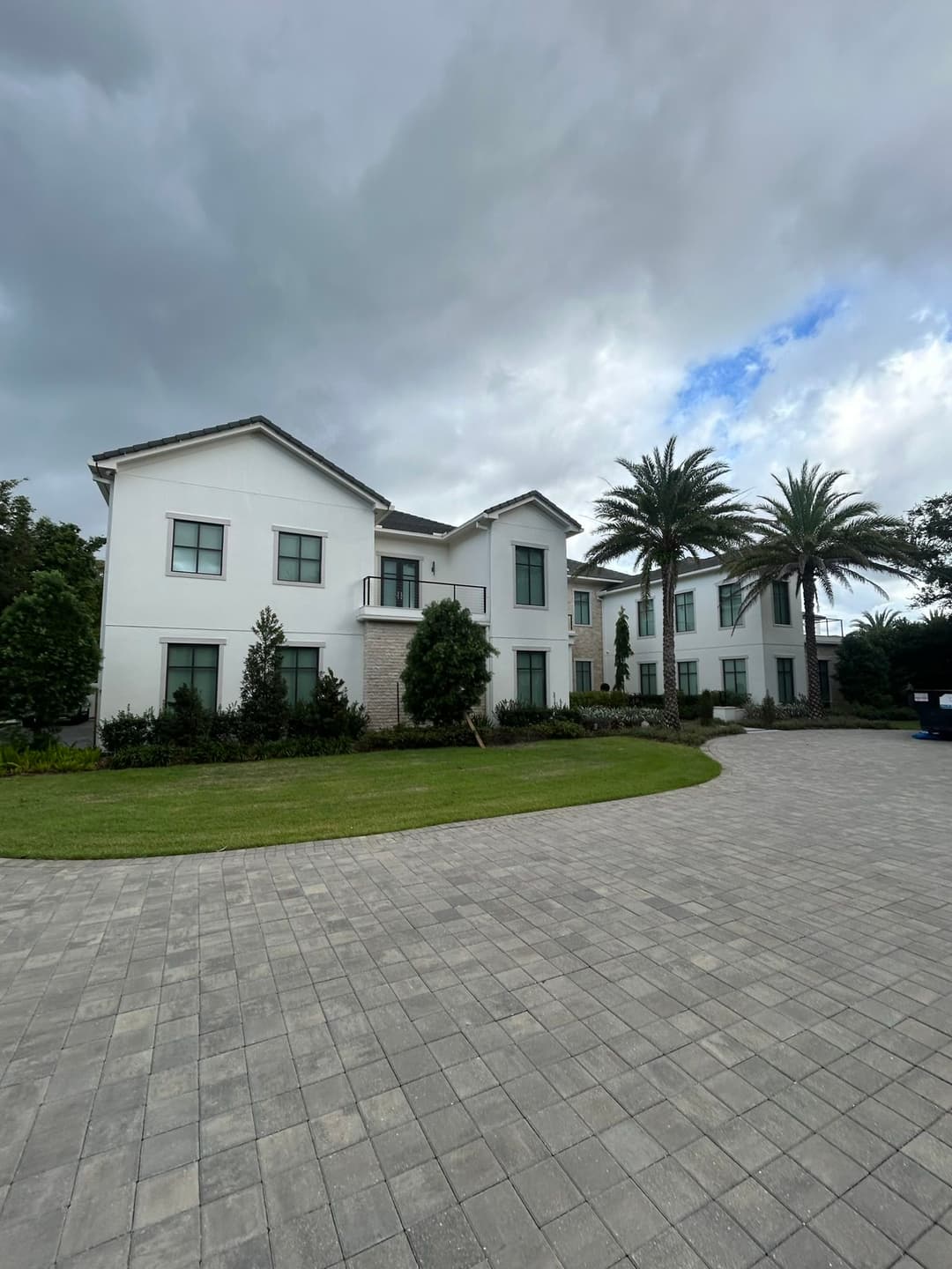 Modern two-story house with palm trees and a landscaped yard in a cloudy backdrop.